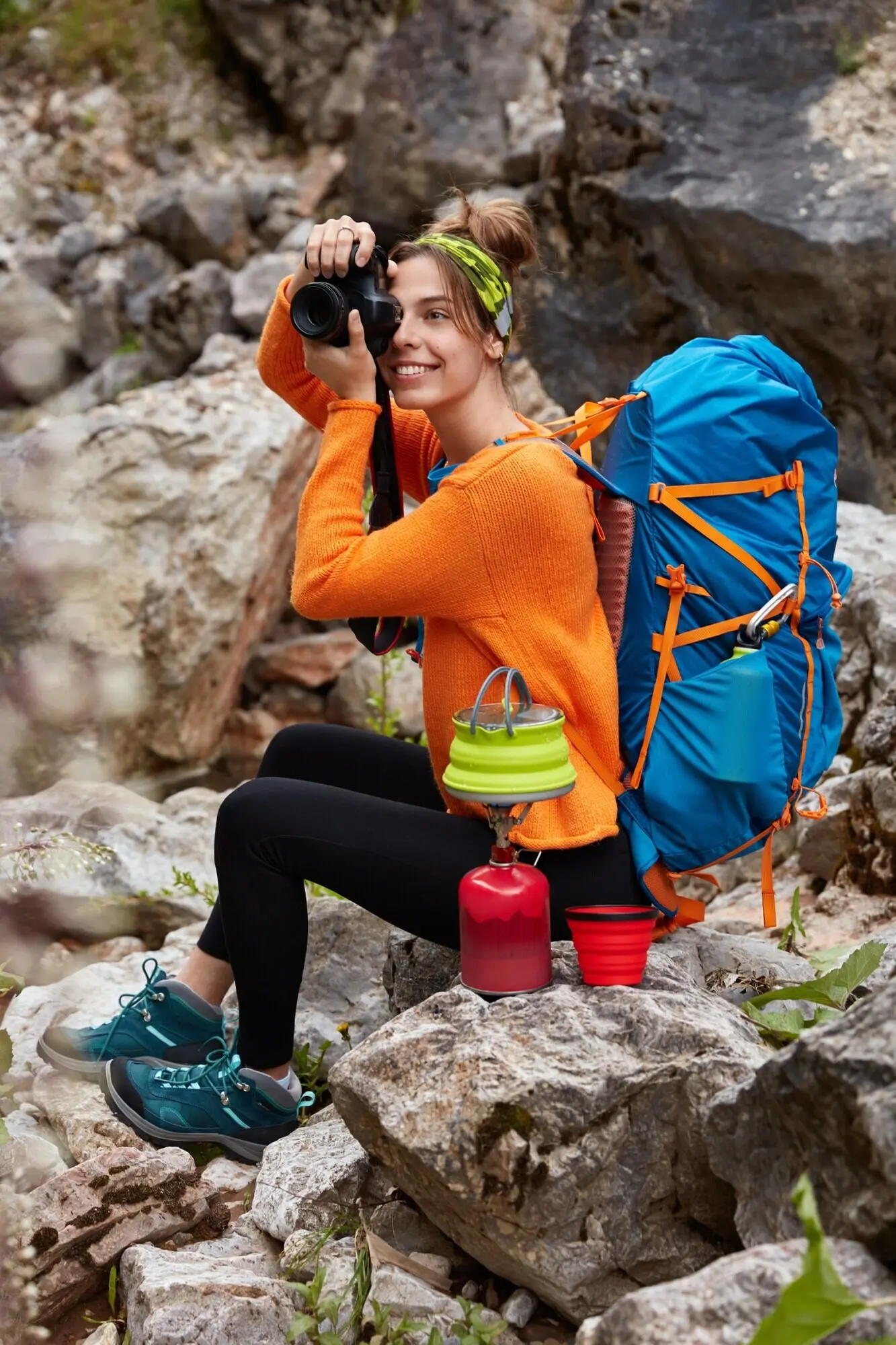 Hochformataufnahme eines fröhlichen Reisenden, der auf Felsen sitzt, mit der Kamera wunderbare Fotos macht, auf einem Campingkocher Kaffee zubereitet und einen orangefarbenen Pullover trägt.