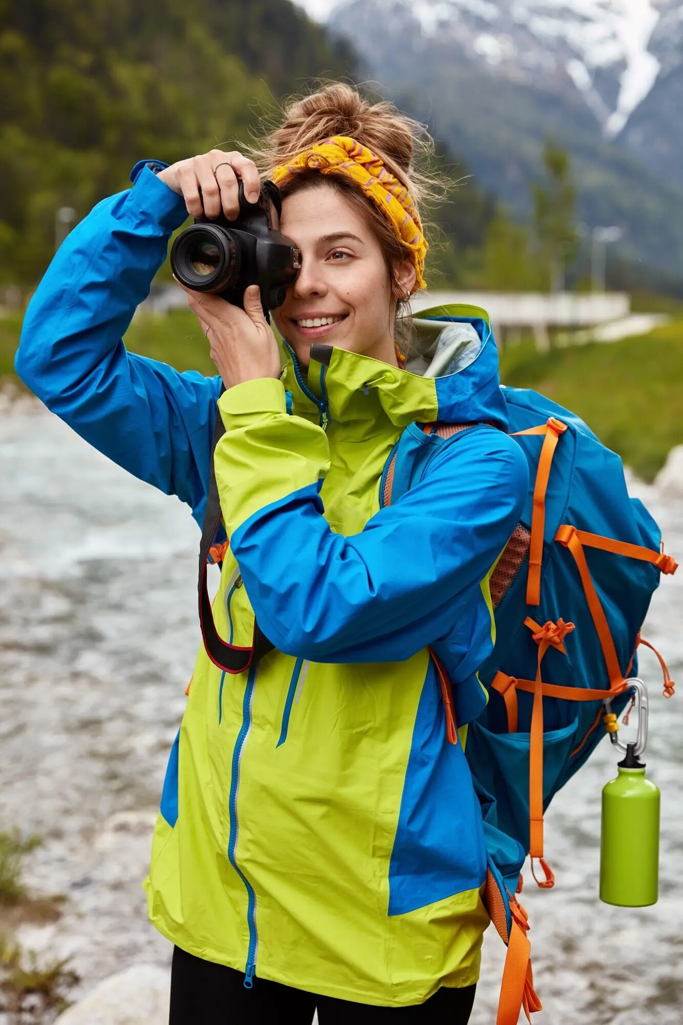 Fröhlicher Tourist posiert vor malerischer Aussicht, trägt einen großen Rucksack, schießt mit der Kamera ein Foto, fotografiert einen Bach und trägt einen Anorak.